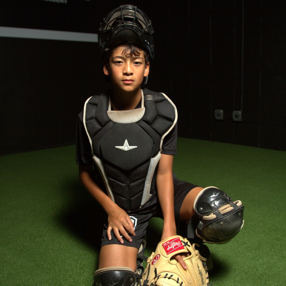 A young baseball catcher in full gear kneels on artificial turf, holding a mitt and wearing a chest protector and helmet.