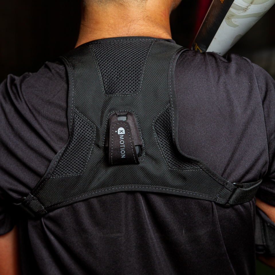 Close-up of a person wearing a black harness with a small electronic device labeled "MOTION" attached to the back. The person is holding a baseball bat over their shoulder.