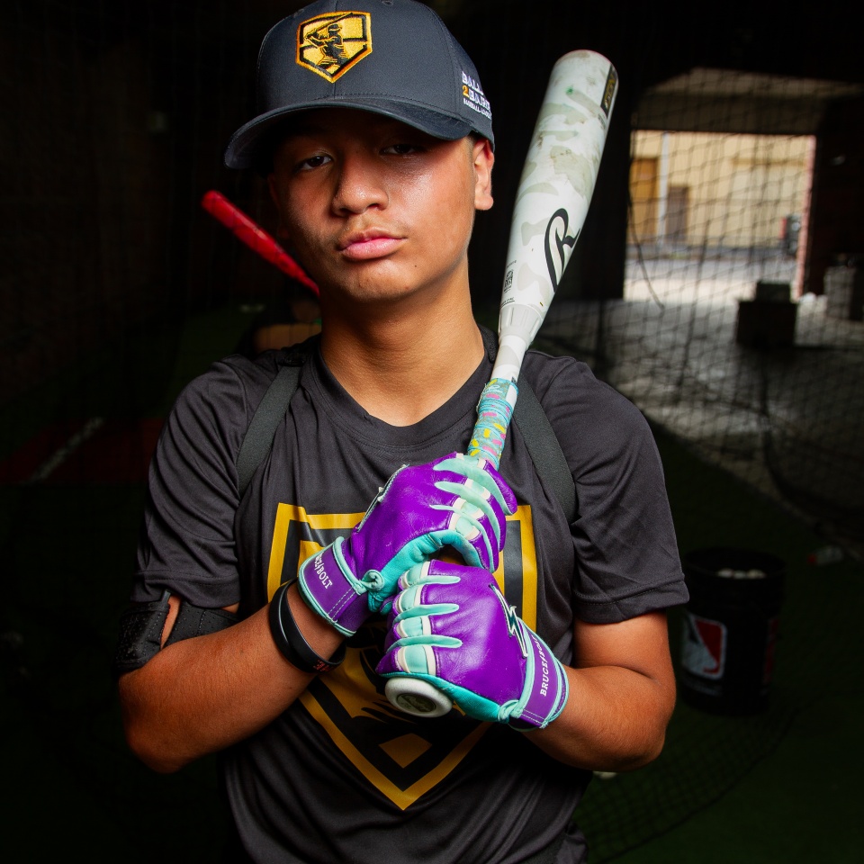 A young baseball player wearing a black cap, black shirt with a yellow logo, and purple gloves, holding a bat over his shoulders inside an indoor batting cage.