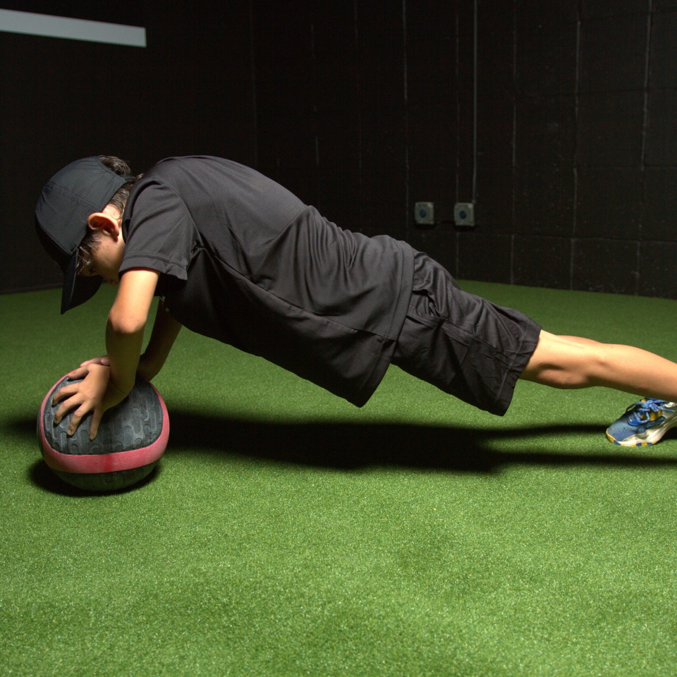 A person wearing black athletic clothing and a cap performs a plank with hands on a medicine ball on artificial turf indoors.