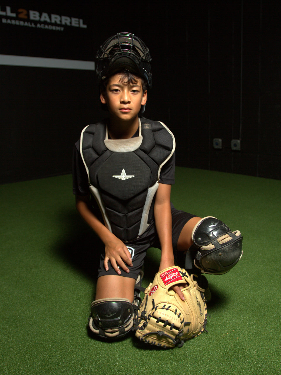 A young baseball catcher in full gear kneels on artificial turf indoors, looking at the camera.