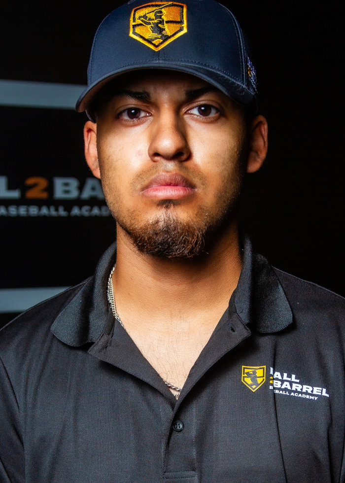 A man in a Ball 2 Barrel Baseball Academy uniform and cap looks at the camera with a neutral expression, standing in front of a branded backdrop.