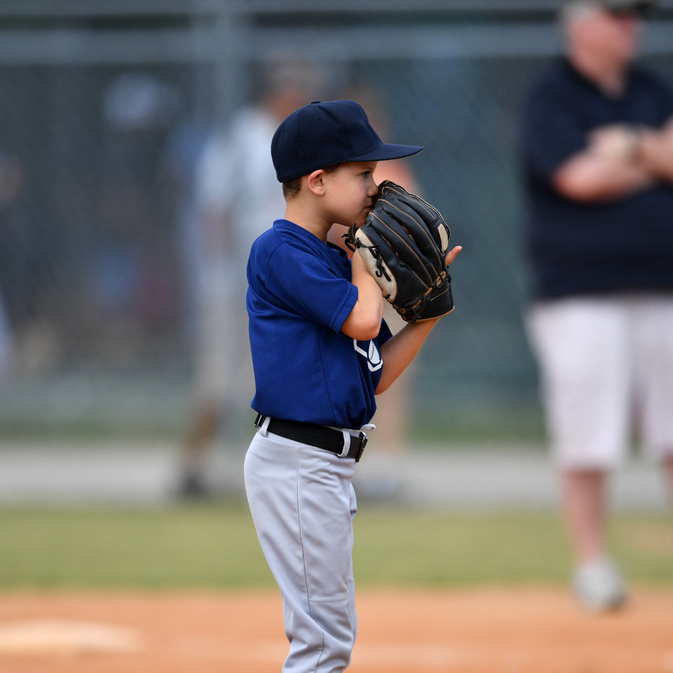 Young boy in a blue baseball uniform and cap stands on a baseball field, holding a glove up to his face, preparing to pitch.