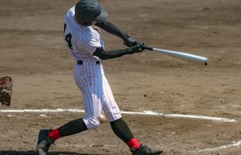 A baseball player in a striped uniform and black helmet swings a bat during a game, making contact with the ball on a dirt field.