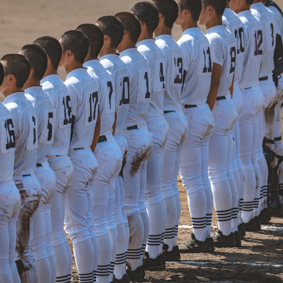 A row of baseball players in white uniforms with numbers on their backs stand side by side on a dirt field, facing away from the camera.