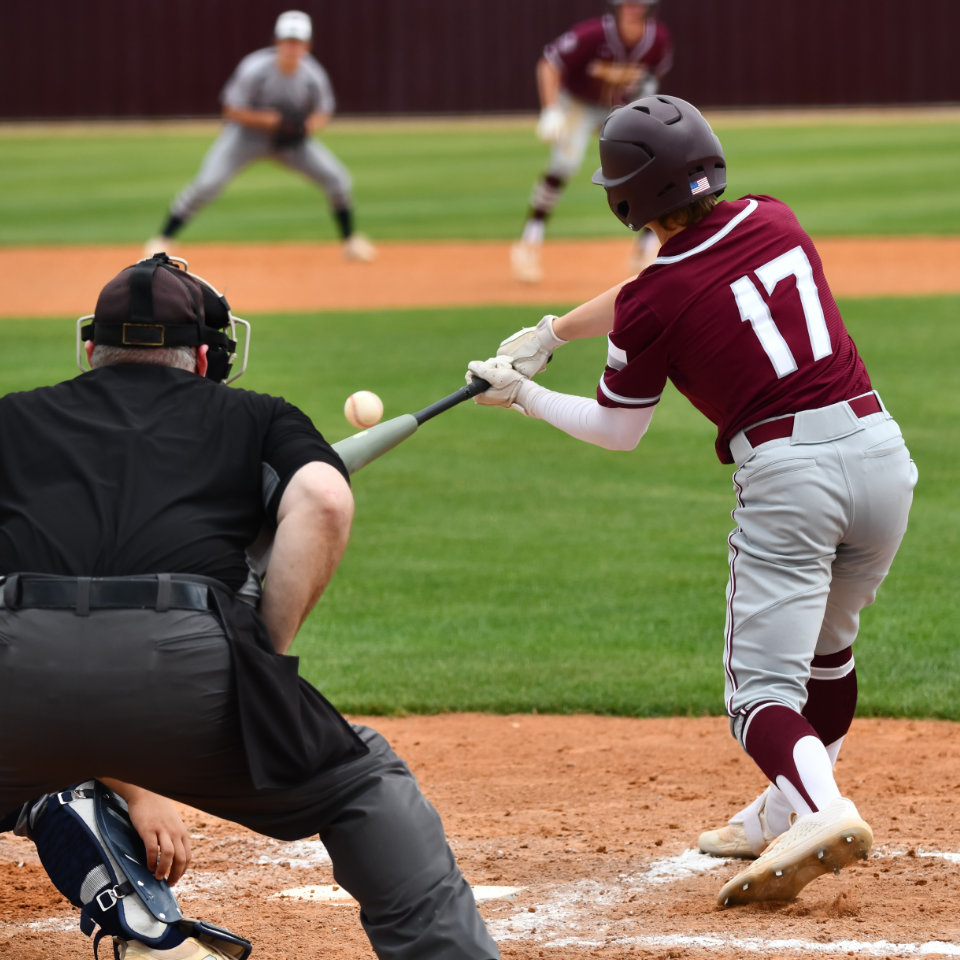 Baseball player wearing number 17 swings at a pitch during a game, with umpire and catcher positioned behind home plate and fielders in the background.