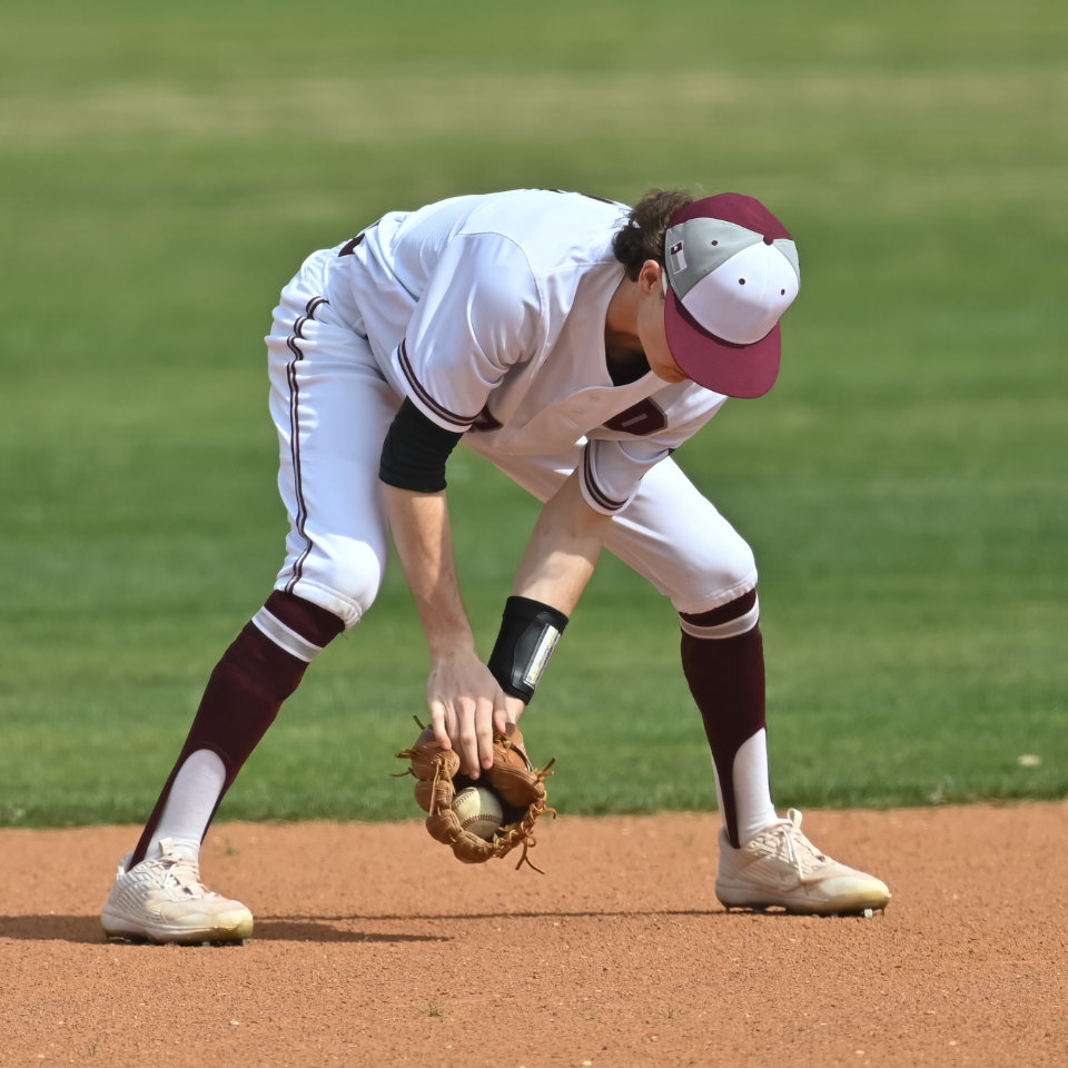 A baseball player in a white uniform and maroon cap fields a ground ball on the infield dirt.