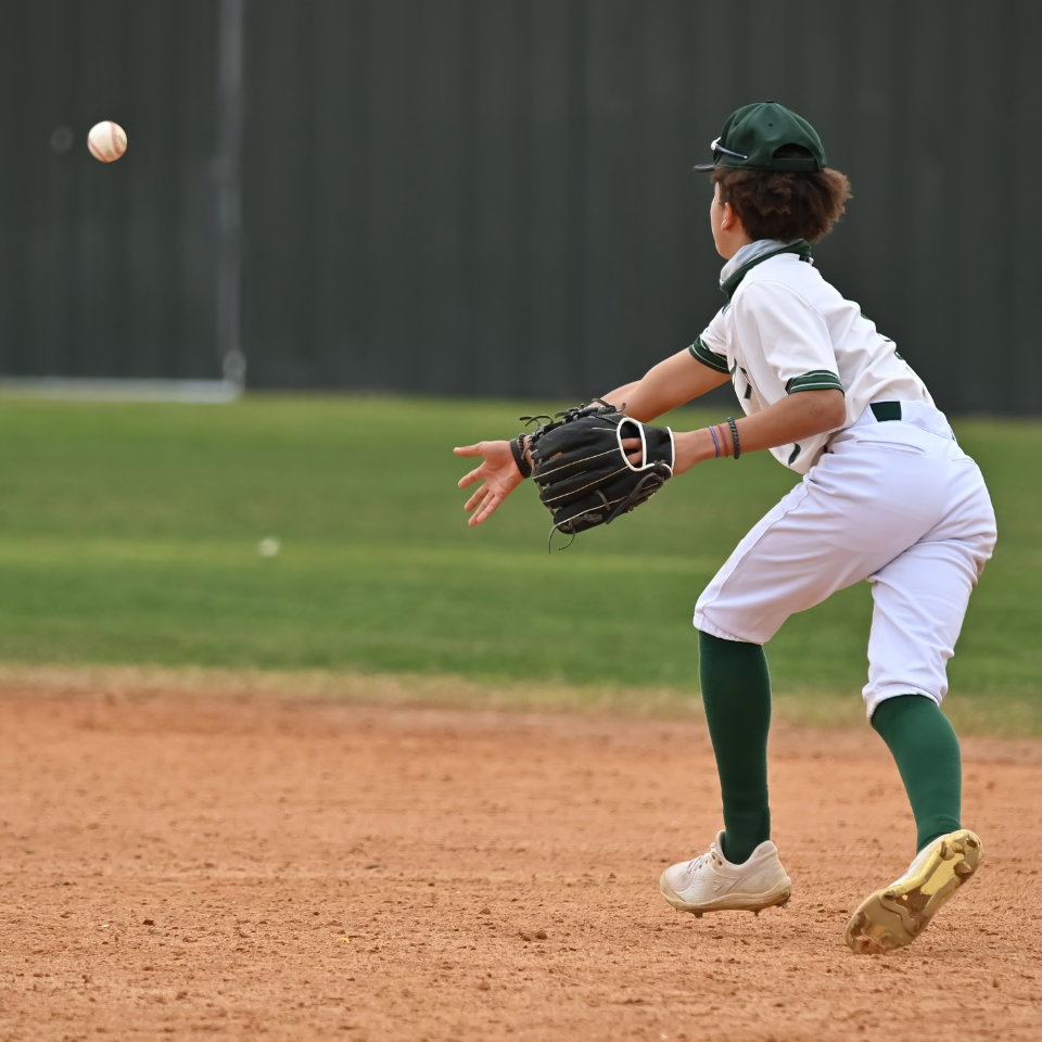 A young baseball player in uniform stands ready with a glove to catch an incoming baseball on a dirt infield.