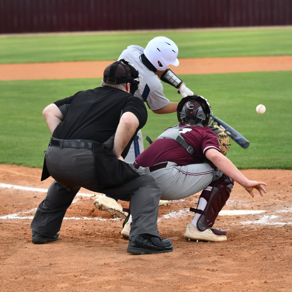 A baseball batter swings at a pitch as the catcher and umpire watch behind him on a dirt baseball field.