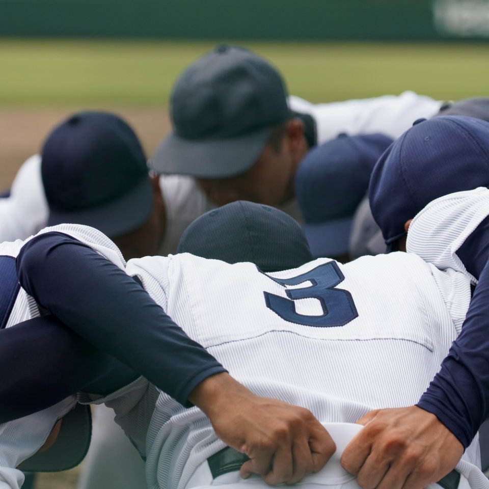 A group of baseball players in uniforms and caps huddle together on a field, with their arms around each other.