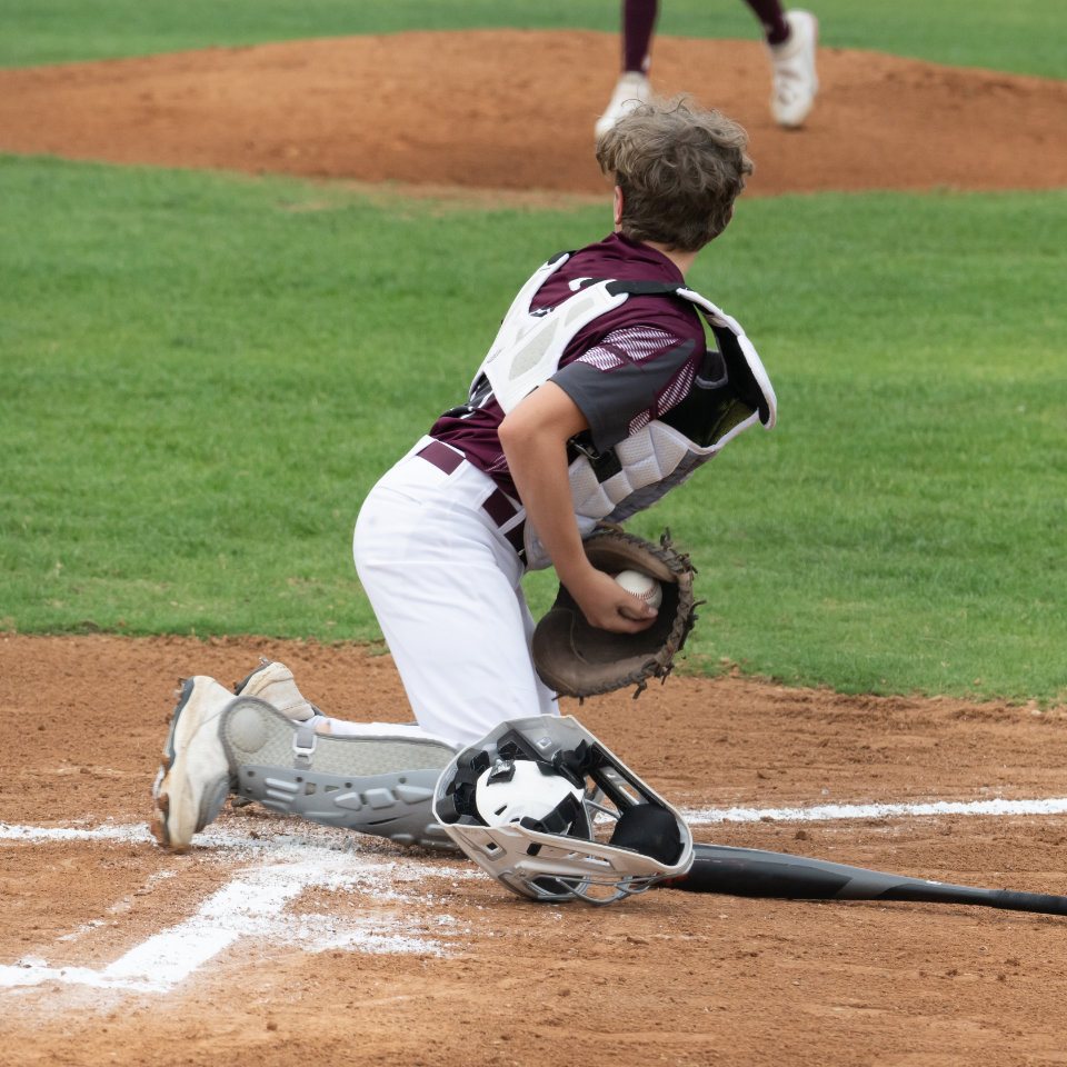 A baseball catcher in maroon and white gear kneels near home plate, holding a glove and ball, with a helmet and bat on the ground.