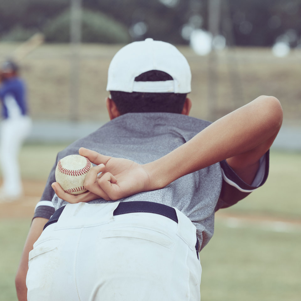 A baseball player in a gray shirt and white cap prepares to pitch, holding a baseball behind his back. Other players and a field are visible in the background.