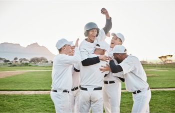 A group of baseball players in white uniforms lift a cheering teammate into the air on a baseball field.