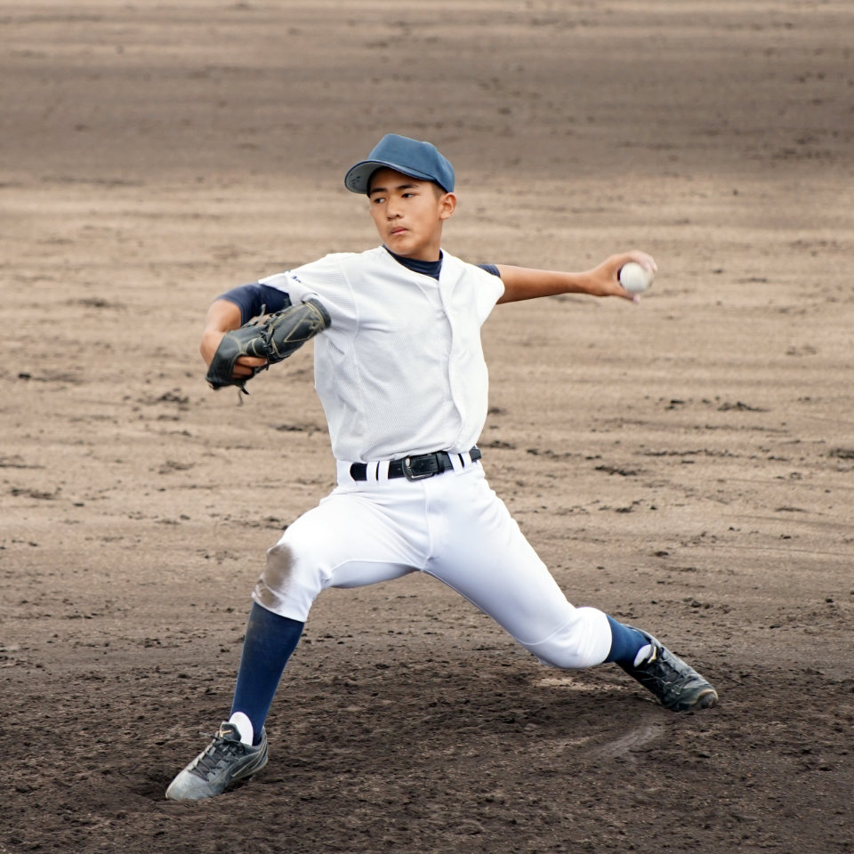 A young baseball player in a white uniform and blue cap is pitching on a dirt field, mid-throw with one leg extended and arm ready to release the ball.