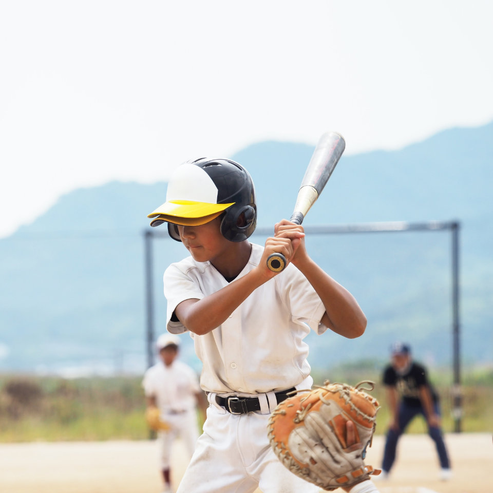 A young baseball player in a white uniform and helmet holds a bat, preparing to hit, with other players and an umpire visible in the background.