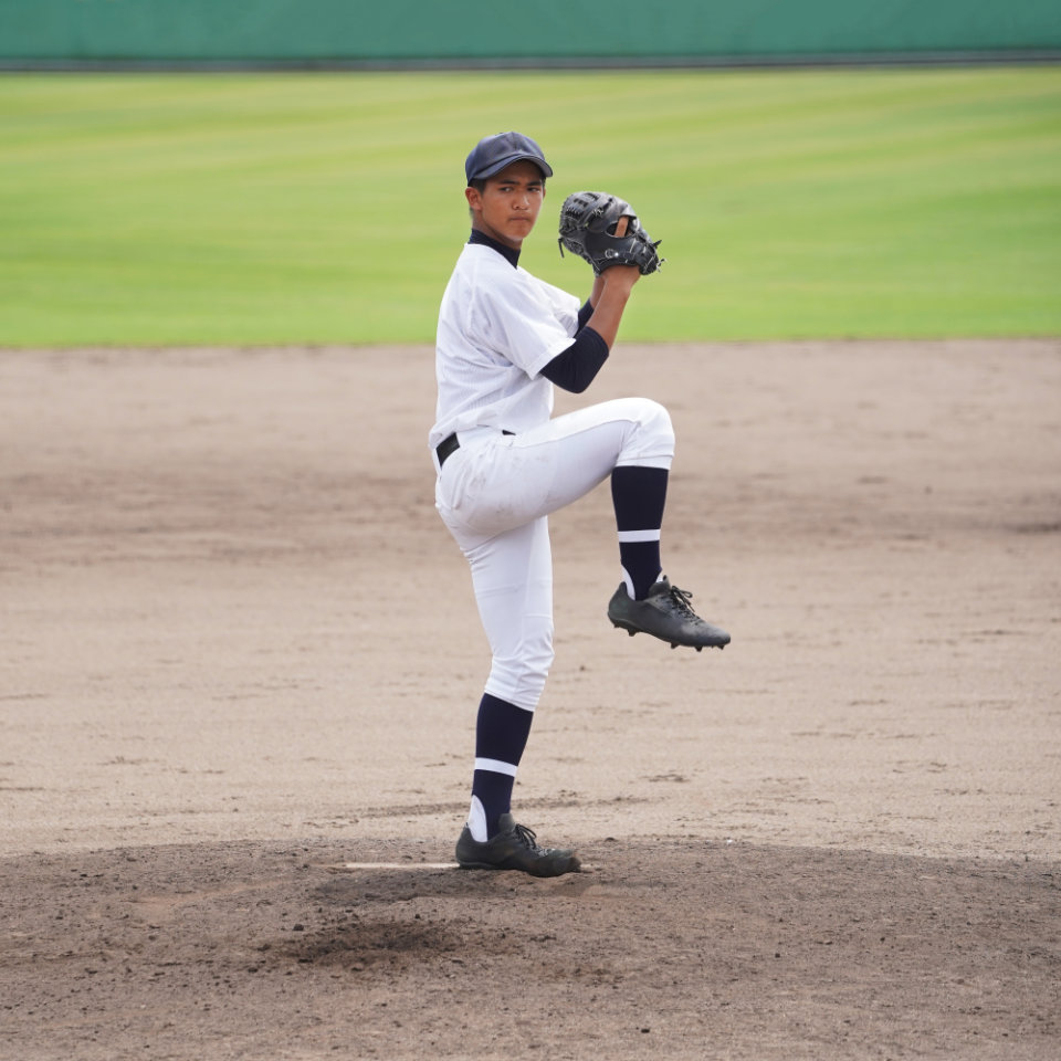 A baseball player in uniform stands on the pitcher's mound, lifting one leg and preparing to throw a pitch on a dirt field with green grass in the background.