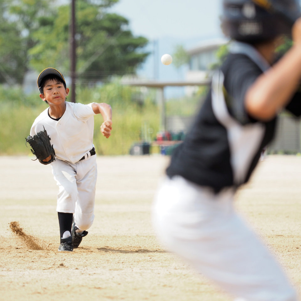 A young baseball player in a white uniform pitches the ball to a batter in black during a game on an outdoor field.