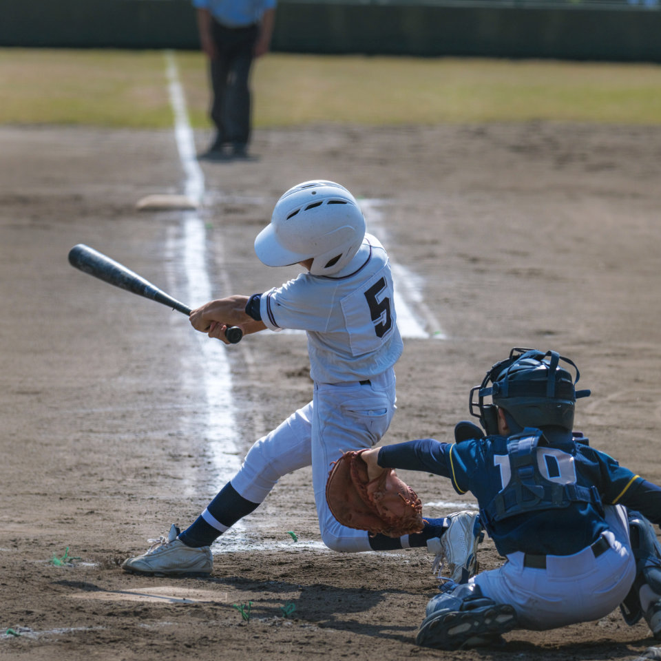 A young baseball player in a white uniform swings a bat at home plate, while a catcher in blue gear crouches behind him on a dirt field.