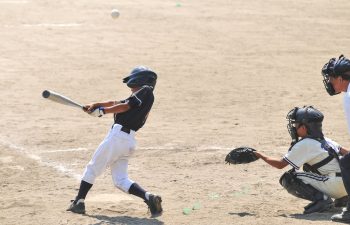 A young baseball player in a black uniform swings at a pitch while the catcher and umpire watch on a dirt field.