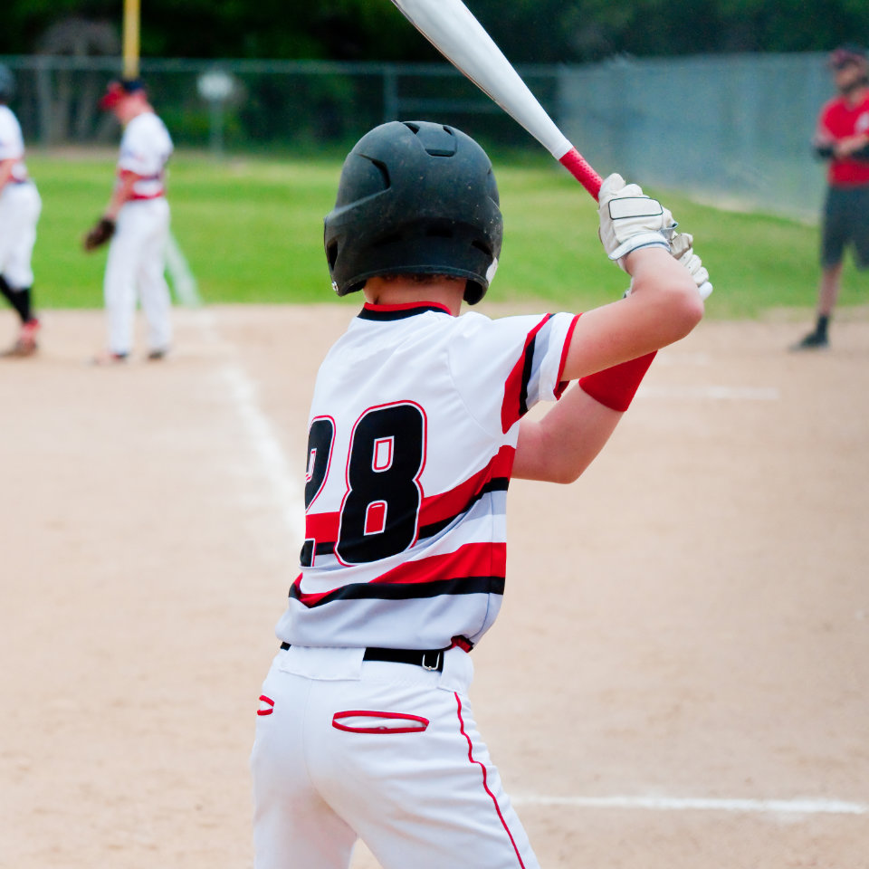 A youth baseball player wearing a helmet and jersey number 28 stands at bat, preparing to hit, with teammates and a coach visible in the background.