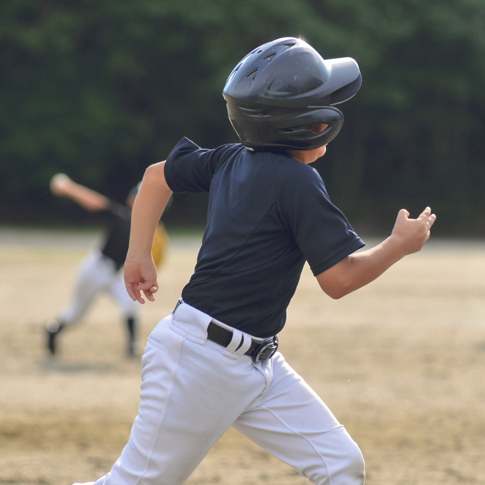 A young baseball player in a black helmet and uniform runs on a dirt field, with another player blurred in the background.