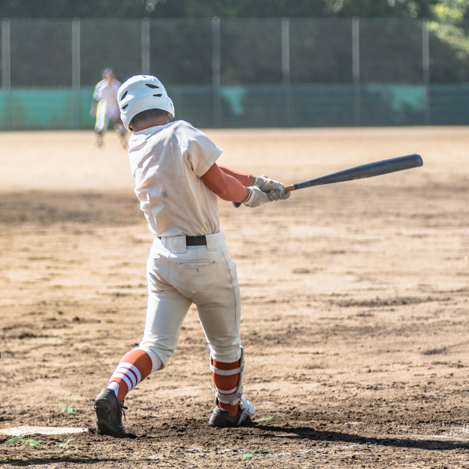 A baseball player in a white uniform and helmet swings a bat during a game on a dirt field, with another player visible in the outfield.