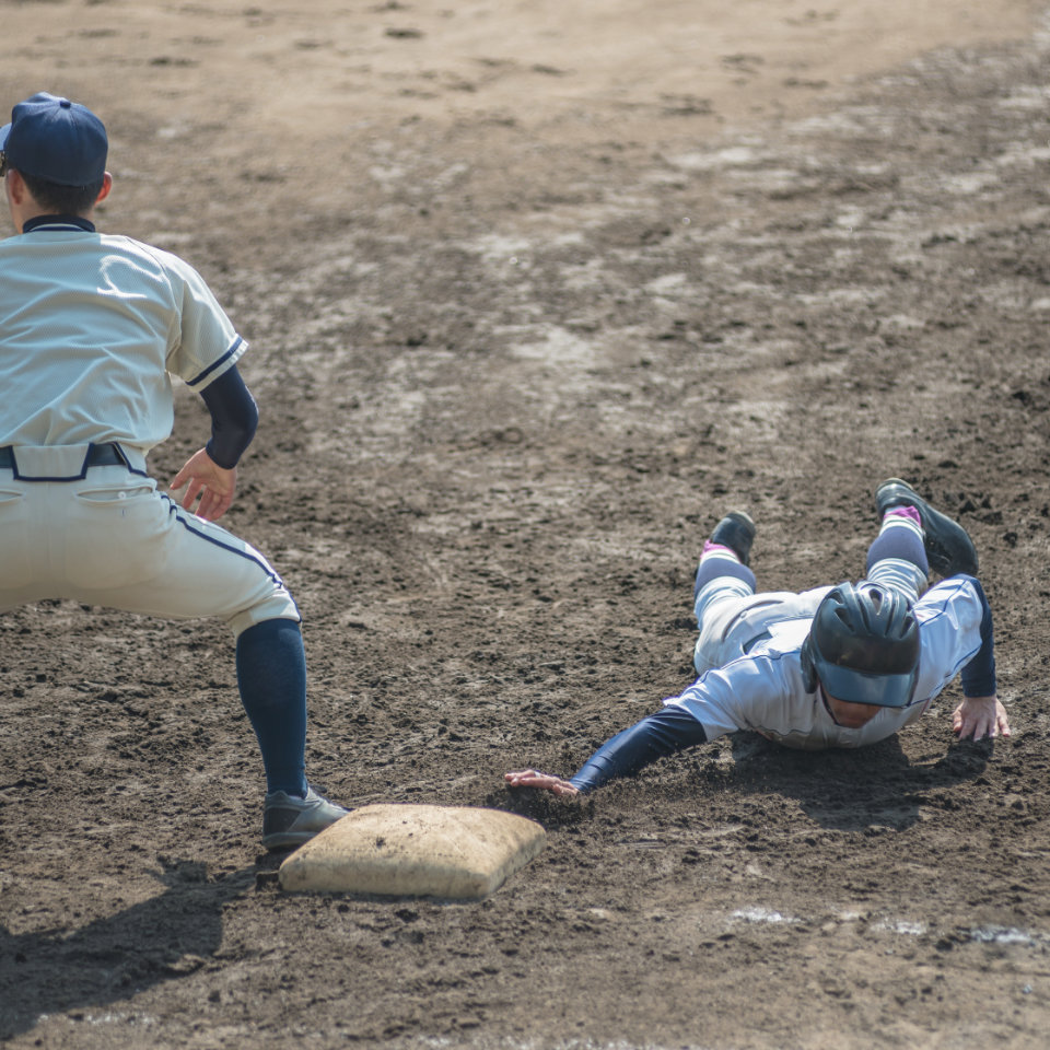 A baseball player slides headfirst toward a base while a fielder in uniform prepares to tag him on a dirt field.
