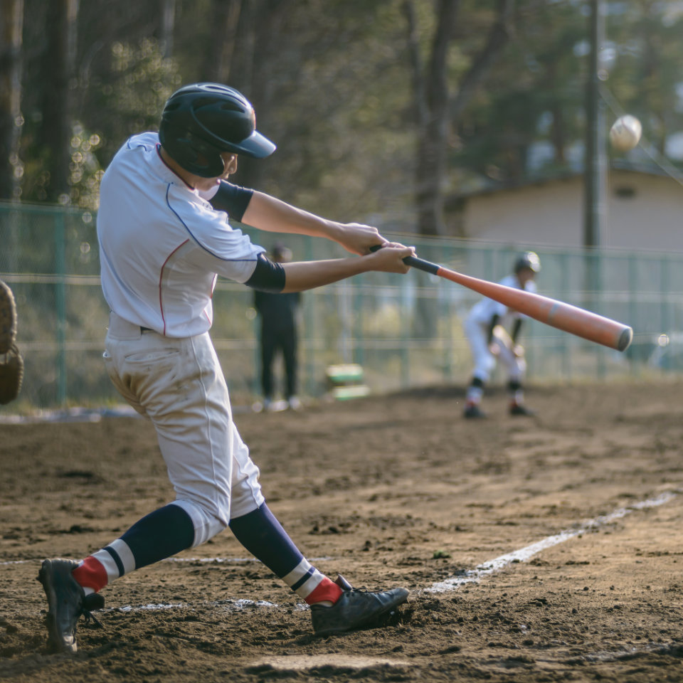 A baseball player in a white uniform swings a bat at a pitch during an outdoor game; another player and an umpire are visible in the background.