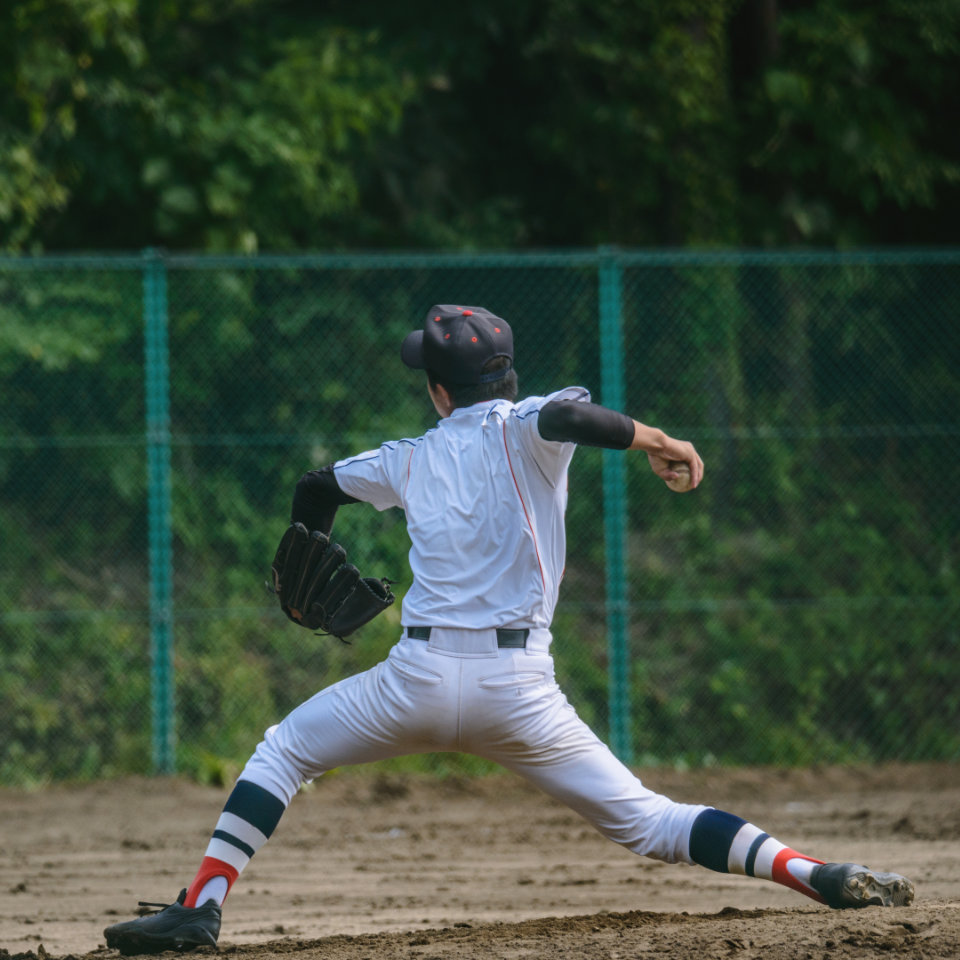 Baseball player in white uniform and cap winding up to pitch on a dirt field, with a green fence and trees in the background.