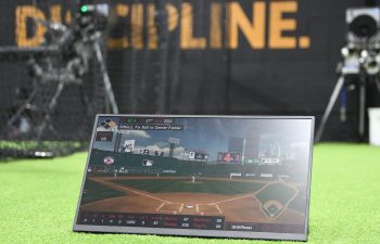 A portable screen displays a baseball game simulation on artificial turf, with training equipment and the word "DISCIPLINE" visible in the background.