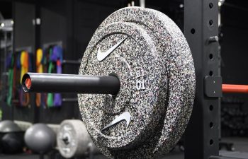 A close-up of a barbell loaded with speckled 10 kg weight plates on a squat rack in a gym, with exercise balls and resistance bands in the background.