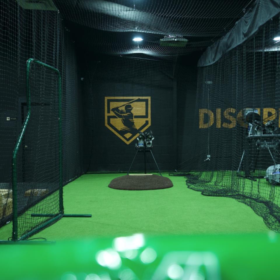 Indoor batting cage with a pitching machine on artificial turf, surrounded by safety nets; a large baseball player logo is visible on the back wall.