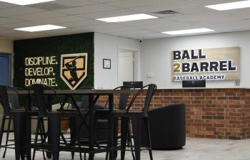 A modern indoor space with high-top tables and chairs, a brick counter, and wall signs reading "DISCIPLINE. DEVELOP. DOMINATE." and "BALL 2 BARREL BASEBALL ACADEMY.