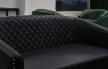 A black quilted leather sofa in a modern lounge area with green chairs, a window, and neon wall signs in the background.