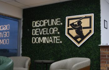 Two grey chairs and a small table sit in front of a green wall with a shield logo and illuminated text reading "DISCIPLINE. DEVELOP. DOMINATE." in an office or lounge area.