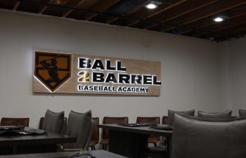 Interior of a room with tables and chairs, featuring a "Ball 2 Barrel Baseball Academy" sign on the wall under exposed wooden ceiling beams.