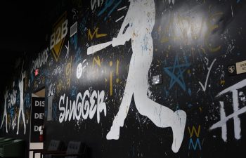 A mural featuring a white silhouette of a baseball batter is surrounded by graffiti-style words and drawings on a black wall.