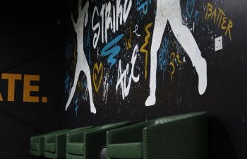 Four green armchairs are lined up along a wall featuring colorful, graffiti-style baseball artwork in a dimly lit room.