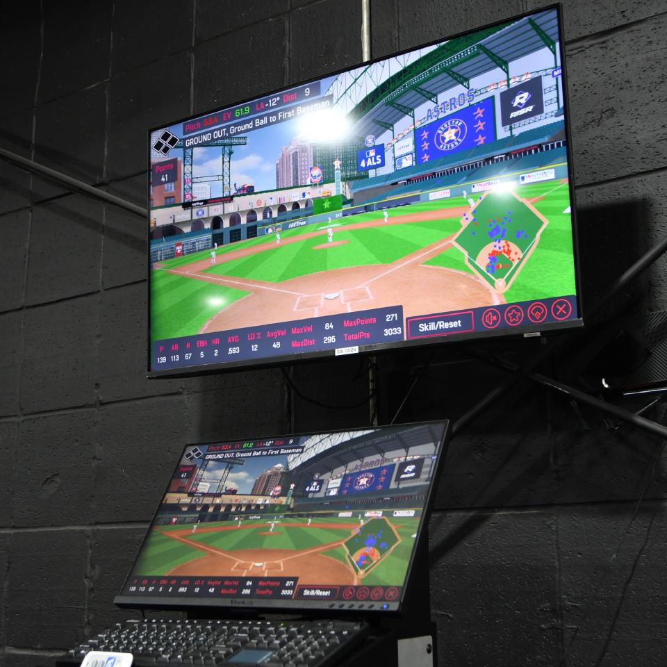 Two screens display a baseball video game simulation with an in-game stadium view and player statistics, set against a black brick wall.
