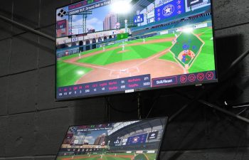 A laptop and monitor display a baseball video game simulation with detailed graphics and stats, set against a black brick wall.
