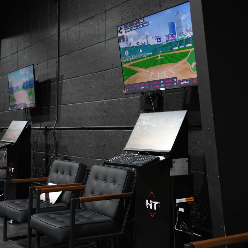 Two black chairs sit in front of a kiosk with a keyboard and two monitors displaying a digital baseball game in a dark room with black walls.