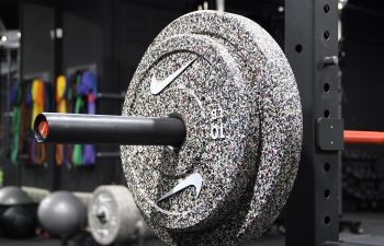 A close-up of a barbell loaded with speckled weight plates on a squat rack in a gym. Fitness equipment and colorful resistance bands are visible in the background.