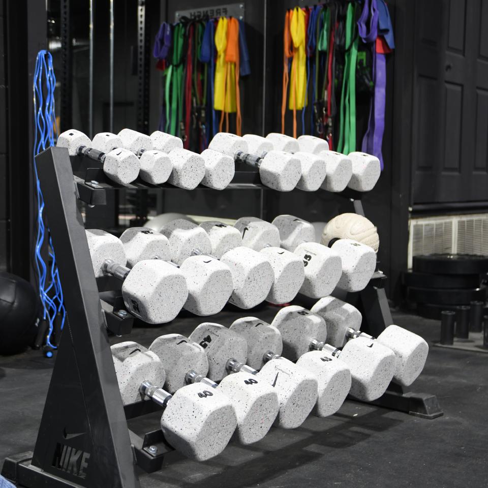 A rack holding three rows of gray hexagonal dumbbells is in a gym. Colorful resistance bands hang in the background.