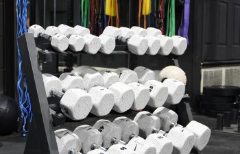 A rack of white hexagonal dumbbells in a gym, with colorful resistance bands hanging on the wall in the background.