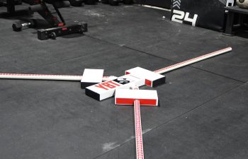 Measurement rods and boxes arranged in an X shape on a gym floor, with weights, benches, and plyometric boxes in the background.
