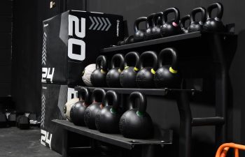 A set of kettlebells on a rack, soft plyo boxes labeled 20 and 24 inches, and orange resistance bands in a gym with black walls and flooring.