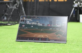 A monitor displaying baseball analytics sits on artificial turf in an indoor training facility with "DISCIPLINE" written on the back wall.