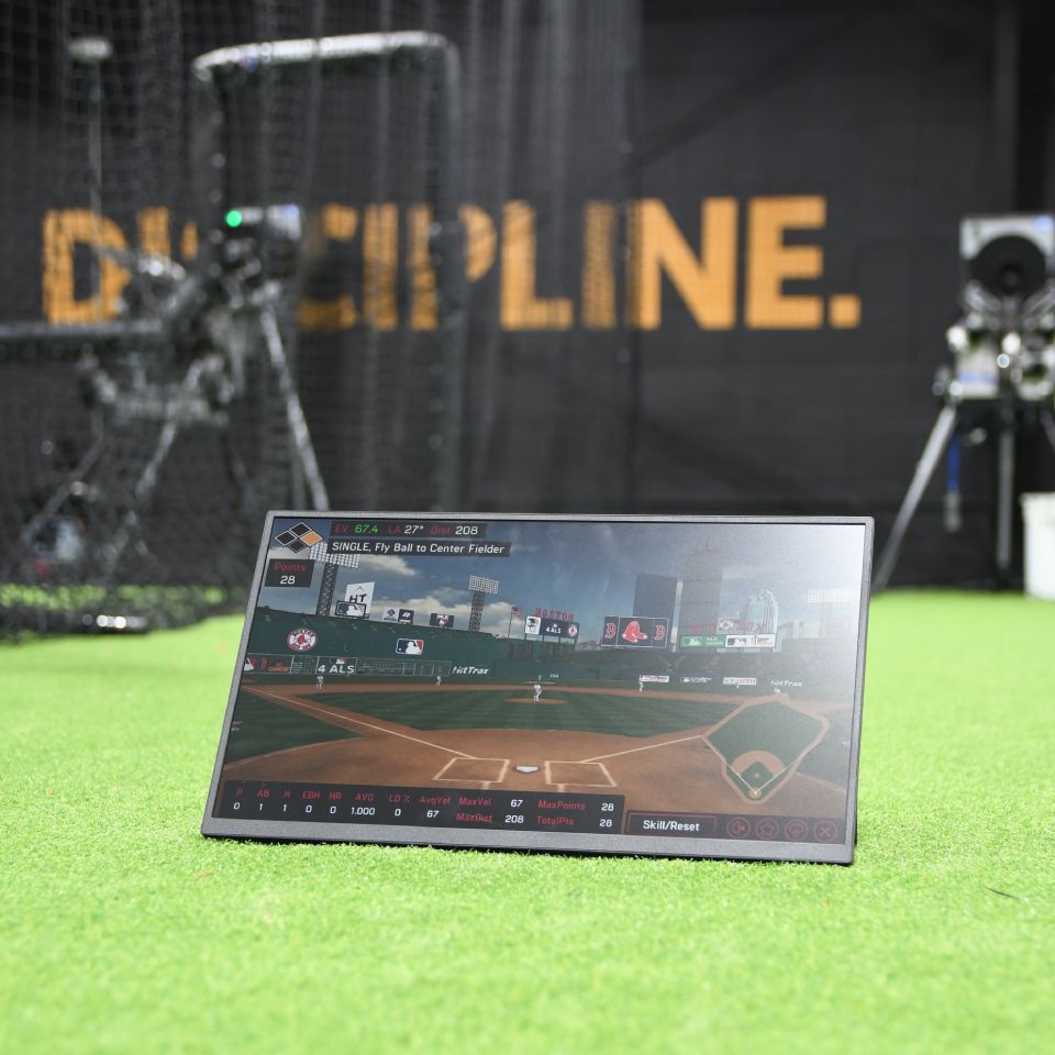 A digital screen displaying baseball game statistics sits on artificial turf in an indoor training facility with "DISCIPLINE" written on the back wall.