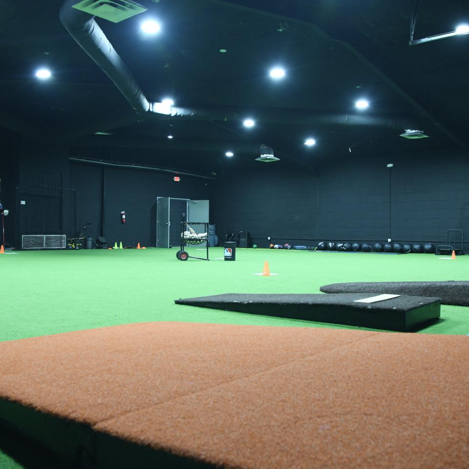 Indoor sports training facility with artificial turf, pitching mound, orange cones, exercise equipment, and netted area in the background.