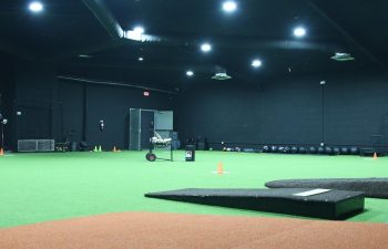 Indoor sports training facility with artificial turf, pitching mound, rubber mat, cones, and equipment under overhead lighting.